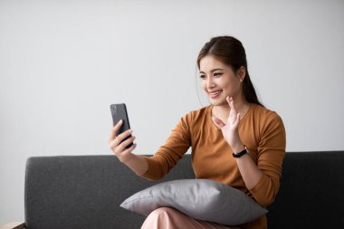 Genç bir kadın koltukta rahatça oturur, gülümser ve akıllı telefonuna el sallar. Rahatlamış ve modern bir ev ortamında bir anlık bağlantının tadını çıkarıyor..