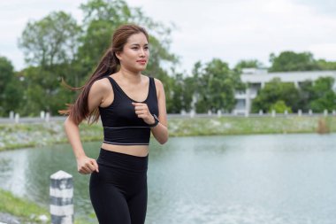 Yeşilliklerle çevrili, göl kenarında koşan atletik giysili genç bir kadın. Spor, sağlık ve açık hava aktiviteleri için mükemmel bir tema..