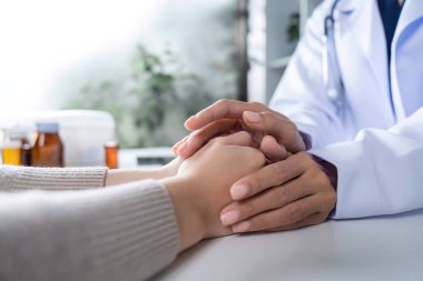 Kadın bir doktor, ellerini tutarak hastaya destek ve cesaret verir. Sağlık hizmetleri ve empati sembolize eder..