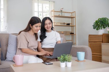 Yaşlı bir anne ve kızı evde birlikte vakit geçiriyorlar, gülümsüyorlar ve rahat bir oturma odasında dizüstü bilgisayar kullanıyorlar..