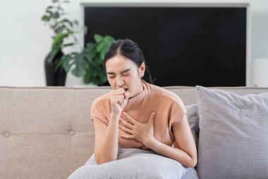 Kanepede oturan bir kadın, öksürüyor ve göğsünü tutuyor, rahatsızlık ya da hastalık belirtileri gösteriyor..