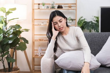 Kafasını tutarak kanepede oturan bir kadın, baş ağrısına işaret ediyor, modern bir oturma odasında, arka planda ahşap bir raf ve iç mekan bitkileri ile.