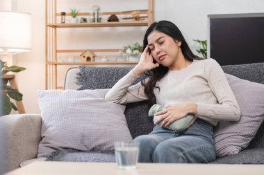 Karın ağrısı ve regl döneminde baş ağrısı olan genç bir kadın elinde ısıtma yastığıyla kanepede oturuyor..