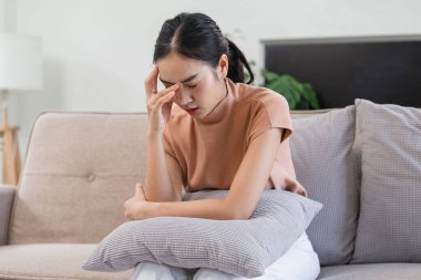 Kanepede oturan genç bir kadın, baş ağrısı ile hasta hissediyor, rahat etmek için yastığını tutuyor, evde rahatsızlık ve acıyı ifade ediyor..