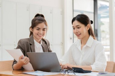 Modern bir ofiste birlikte çalışan iki genç iş kadını, bir dizüstü bilgisayar ve belgeler kullanarak, gülümseyerek ve nişanlanarak,.