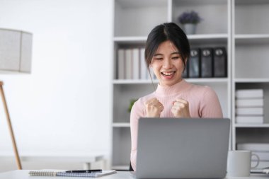 Genç bir kız öğrenci, modern bir ev ofisinde online olarak çalışırken mükemmel sınav sonuçlarını kutluyor ve e-öğrenme ve akademik başarının sevincini gösteriyor..