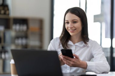 Çağdaş bir ofis ortamındaki masasında dizüstü bilgisayar ve akıllı telefon ile birden fazla iş yapan profesyonel bir iş kadını..