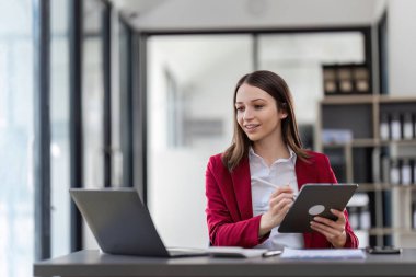 Çağdaş bir ofis ortamında masasında dizüstü bilgisayar ve tabletle birden fazla iş yapan profesyonel bir iş kadını..