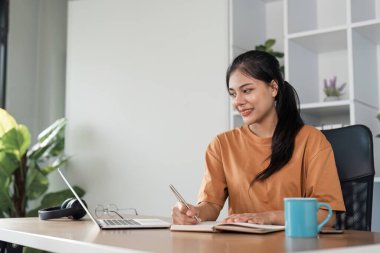 Genç bir kız, bir dizüstü bilgisayar ve defter kullanarak, modern bir ofis ortamında bir masada oturarak online olarak çalışıyor..