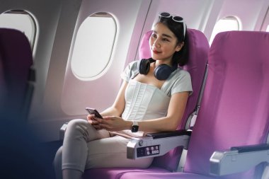 Bir kadın uçmaktan zevk alır, akıllı telefonu ve kulaklıklarıyla rahat bir şekilde oturur, modern seyahat ve uçuş rahatlığı sergiler..