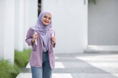 Başörtüsü ve günlük giysiler içindeki genç Müslüman öğrenci çağdaş bir binanın önünde dikilirken rahatça gülümsüyor..