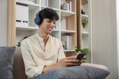 Genç bir adam modern odasında rahat bir şekilde oturur, koltukta dinlenir, telefonuyla oynar ve kulaklık takar..