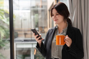 Takım elbiseli kendine güvenen bir iş kadını, akıllı telefonunu modern ofis ortamında kullanır. Profesyonelliğini ve bağlantısını gösterir..