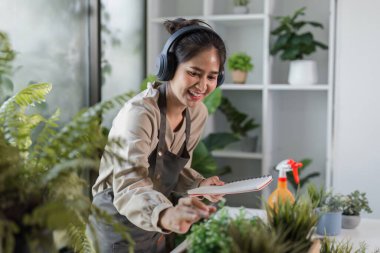 Kulaklık takan bir kadın ev ortamında canlı bitkilerle çevrili iç bahçesiyle ilgilenir..