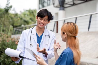 Bir erkek doktor ve bir danışman asistanı, profesyonel sağlık hizmetleri işbirliğini sergileyen çağdaş bir binanın dışında çalışmayı tartışırlar..