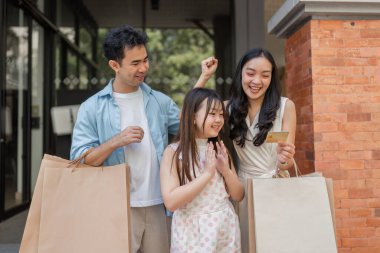 Neşeli bir aile, alışveriş yapmanın heyecanını bir kredi kartıyla yaşıyor, modern bir şehir ortamında mutluluk ve beraberlik sergiliyor..