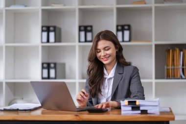 Profesyonel iş kadını, organize dosyalarla çevrili bir dizüstü bilgisayar ve hesap makinesi kullanarak ofis masasında çalışmaya odaklanmış..