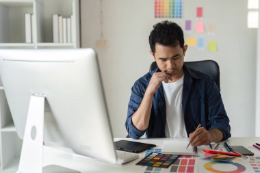 Genç bir erkek grafik tasarımcısı modern ofis ortamında bilgisayar ve renk örnekleri kullanarak yaratıcı bir proje üzerinde çalışıyor..