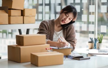 Evde çalışan genç bir kadın, online satış işini yönetiyor. Paketlerle çevrili ve emirleri koordine etmek için dizüstü bilgisayar ve telefon kullanıyor..