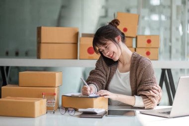 Evde çalışan, internetten ürün satan genç bir kadın. Etrafı kutularla çevrili, e-ticaret işi için bir laptop ve telefon kullanıyor..