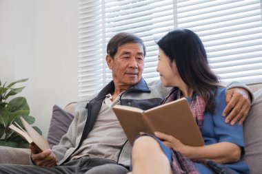 Yaşlı bir çift modern bir oturma odasında dinleniyor, kitap okuyor ve birlikte kaliteli vakit geçiriyorlar. Sıcak ev ortamı ve huzurlu anlar.