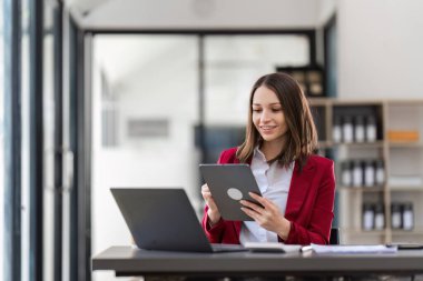 Çağdaş bir ofis ortamında masasında dizüstü bilgisayar ve tabletle birden fazla iş yapan profesyonel bir iş kadını..