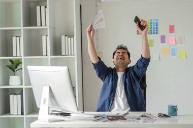 Neşeli bir grafik tasarımcısı, renk örnekleri ve dijital araçlarla çevrili modern bir ofiste başarılı bir projeyi kutluyor.