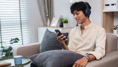 Genç bir adam modern bir oturma odasında rahat bir şekilde oturuyor, kulaklık takıyor ve sosyal medyayı keşfetmek için akıllı telefonunu kullanıyor..