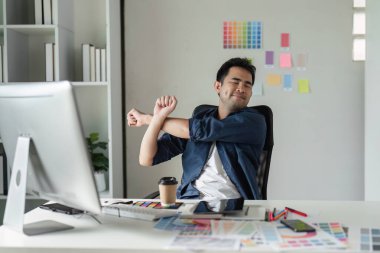 Grafik tasarımcısı, tasarım araçları ve renk örnekleriyle çevrili modern bir ofiste, sağlıklı bir iş-yaşam dengesini teşvik ederek esneyip gevşetiyor..