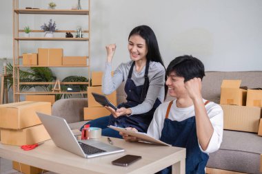 Neşeli bir çift, posta kutuları, dizüstü bilgisayarlar ve tabletlerle çevrili ev ofislerinde başarılı online satışlarını kutluyor..