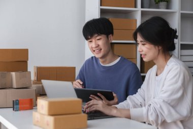 Çevrelerinde kargo kutuları ve bir dizüstü bilgisayar ve tablet üzerinde çalışan genç bir çift evlerinden online olarak küçük işletmelerini yönetiyorlar..