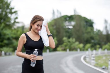 Atletik giyinen genç bir kadın parkta egzersiz yapıyor, elinde bir su şişesi ve havlu tutuyor, açık hava spor yaparken susuz ve sağlıklı kalıyor..