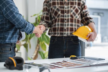 Modern bir ofiste iki erkek mühendis el sıkışıyor, planlar ve güvenlik teçhizatlarıyla bir inşaat projesinde işbirliği yapıyor..