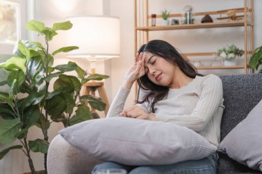 Kanepede oturan bir kadın başını acıdan tutarak, rahat bir oturma odasında bitkilerle ve hafif ışıkla baş ağrısından muzdarip..