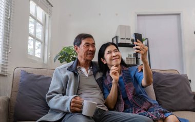 Mutlu bir emekli çift kanepede oturuyor, kahvenin tadını çıkarıyor ve sıcak bir evde selfie çekiyorlar..