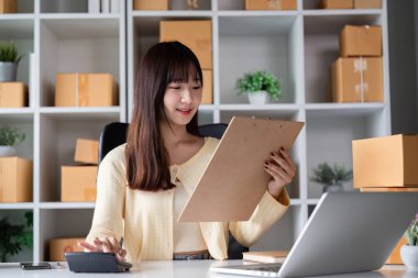Modern bir ev ofisinde çalışan genç bir bayan girişimci, bir dizüstü bilgisayar, dosya ve kutularla çevrimiçi satış yönetiyor..