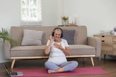 Elderly woman wearing headphones, sitting on a yoga mat, enjoying relaxing music in a modern living room setting. clipart