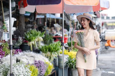 Genç bir kadın hareketli bir şehir pazarında elinde bir buket çiçekle gezmekten ve renkli şemsiyeler altında gülümsemekten zevk alır..