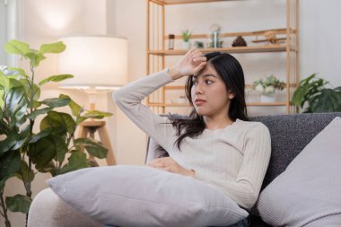 Evdeki bir kanepede oturan genç bir kadın, alnını tutuyor ve endişeli görünüyor, kendini kötü ya da hasta hissettiğini gösteriyor..