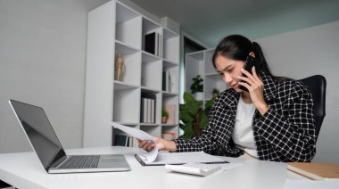 Focused businesswoman working late in a modern office, multitasking with a laptop and phone, showcasing dedication and professionalism. clipart