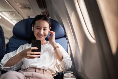 Profesyonel bir iş kadını uçakta rahatlar, uçuş sırasında akıllı telefonunu kullanır, modern seyahat ve iş hayatı dengesini somutlaştırır..