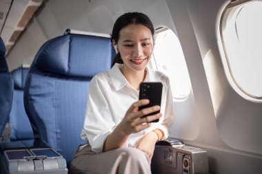 Bir iş kadını uçakta seyahat ederken akıllı telefonunu kullanır, modern çalışma esnekliğini ve uçuş verimliliğini gösterir.