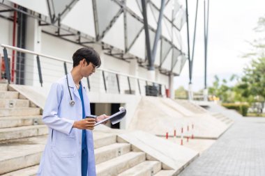 Beyaz önlüklü genç bir doktor modern bir binanın dışındaki hasta vakalarını inceleyerek sağlık ve çağdaş mimarinin bir karışımını sergiliyor..