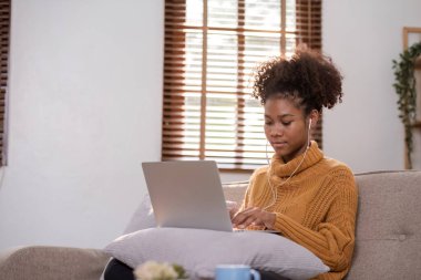 Evde çalışan genç Afrikalı bir kadın, kulaklık takıyor, dizüstü bilgisayar kullanıyor ve rahat bir oturma odasında dinleniyor..