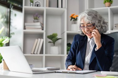 Yaşlı bir iş kadını, bir şirketin sahibi, masasında çalışırken telefonla konuşuyor, liderlik ve profesyonellik gösteriyor..