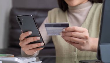 Güzel genç bir kadın internetten alışveriş yapmak için dizüstü bilgisayar kullanıyor. Evden bir şeyler alıyor ve internet bankacılığı uygulaması üzerinden kredi kartıyla ödeme yapıyor..