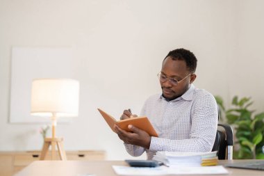 Belgelerle çalışan Afrikalı Amerikalı işadamı, ofiste notlar alıyor, performans analizi raporu hazırlıyor...