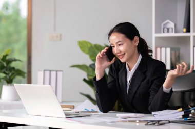 Asian female accountant Discussing business over the phone in an office with work plan documents on the table. clipart