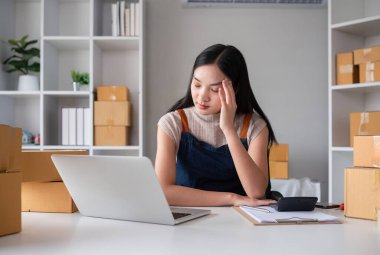 Küçük bir KOBİ işletmesi sahibi, evdeki masasında oturup ürünlerini kutulara doldururken yorgun hissediyor..