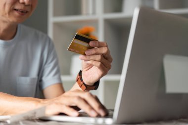Kredi kartı tutan ve online alışveriş için dizüstü bilgisayar kullanan, ev ortamında dijital işlemlerin kolaylığını gösteren odaklanmış yaşlı bir adam..